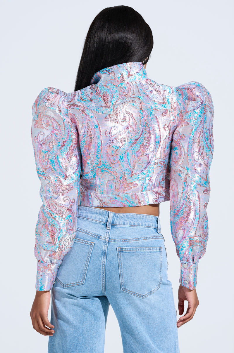 The person is elegantly showcasing the back of their outfit, wearing the DELILAH PUFF SHOULDER BOMBER with a shiny brocade pattern, paired with light blue jeans.