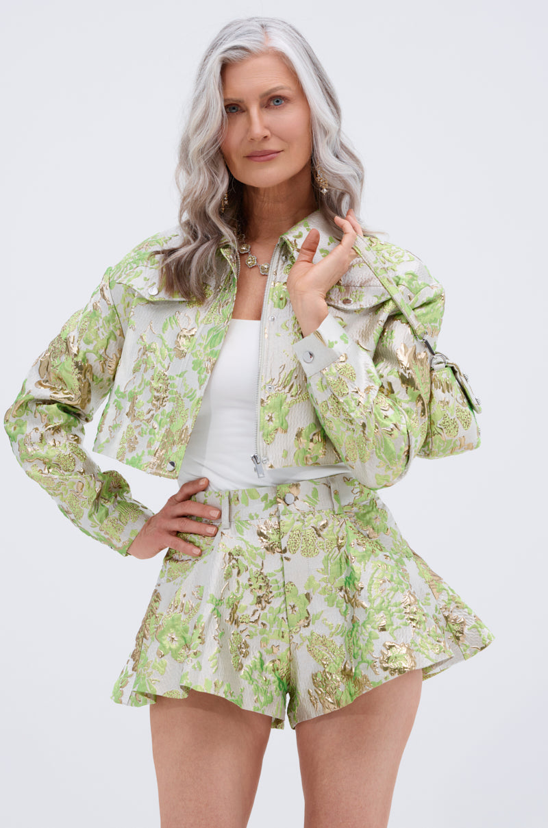 A woman with long, wavy gray hair poses confidently against a plain light background, wearing the HOT SHOT BROCADE BOMBER—an elegant green and gold cropped jacket with metallic floral embroidery—over matching shorts and a white top.