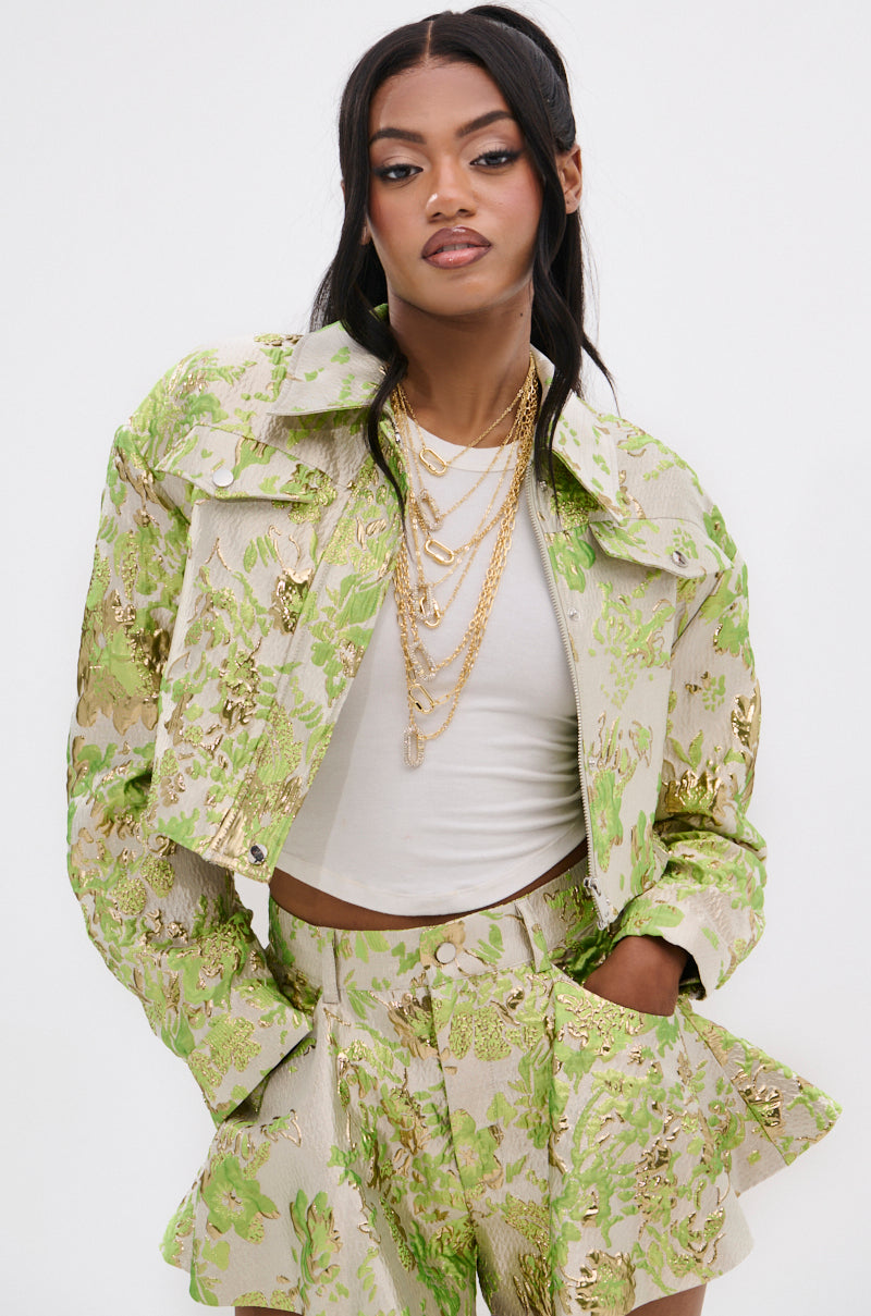 A woman poses confidently in a HOT SHOT BROCADE BOMBER with metallic floral embroidery, matching green and gold high-waisted shorts, a white crop top, and layered gold necklaces against a plain white background.