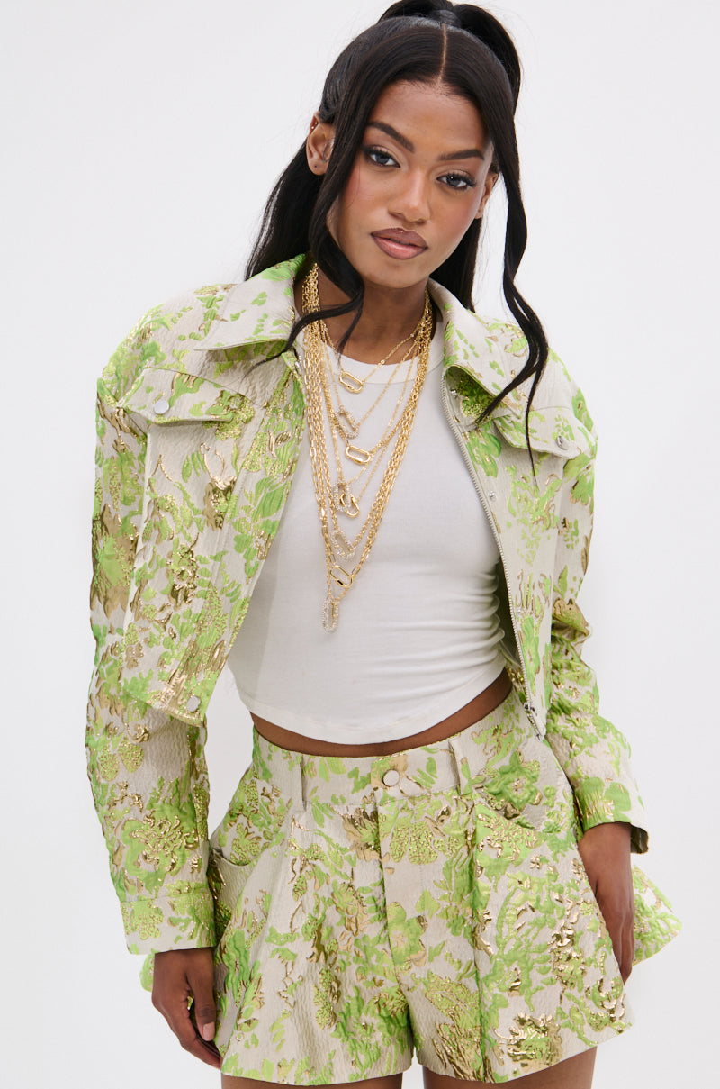 A woman with long dark hair in a high ponytail wears layered gold necklaces, a white cropped top, and the HOT SHOT BROCADE BOMBER with a matching skirt set featuring metallic floral embroidery, standing against a plain light background.