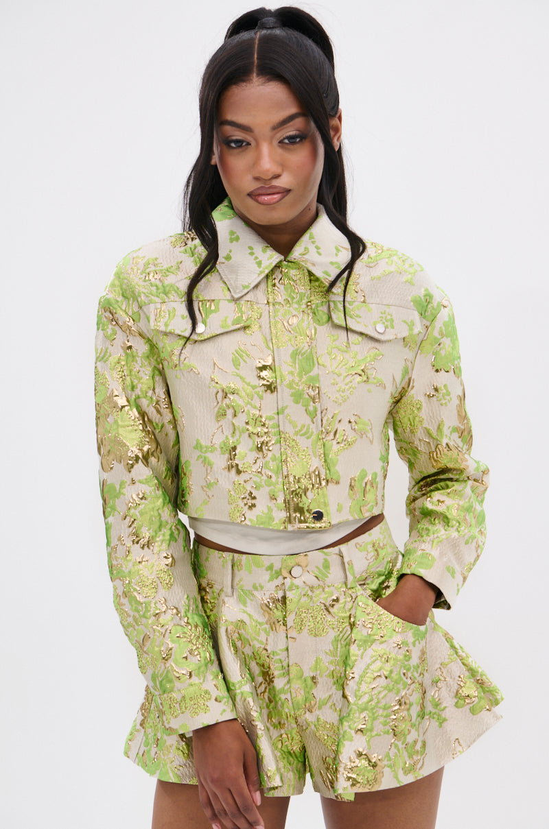 A woman poses in the HOT SHOT BROCADE BOMBER set—cropped long-sleeve jacket and high-waisted shorts in shimmering green, gold, and beige floral brocade—her dark waves contrasting against a plain white background.