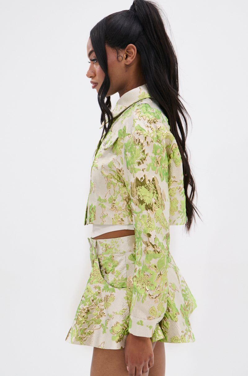 A woman with long dark hair in a high ponytail is shown in profile wearing the HOT SHOT BROCADE BOMBER set with metallic green and gold floral embroidery, against a plain white background.