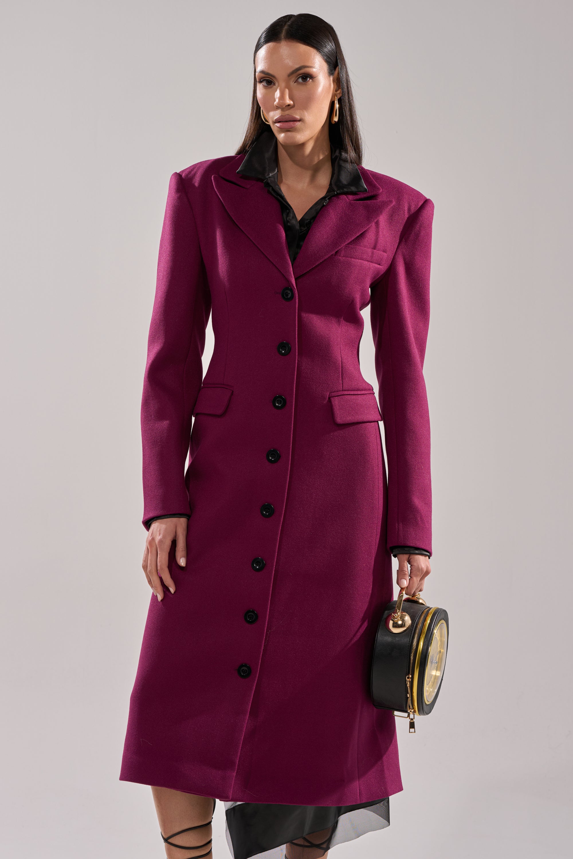 A woman wears the SCARLETT LUXE TRENCH IN PURPLE with black buttons and structured shoulders, holding a round black handbag, standing against a plain light gray background.