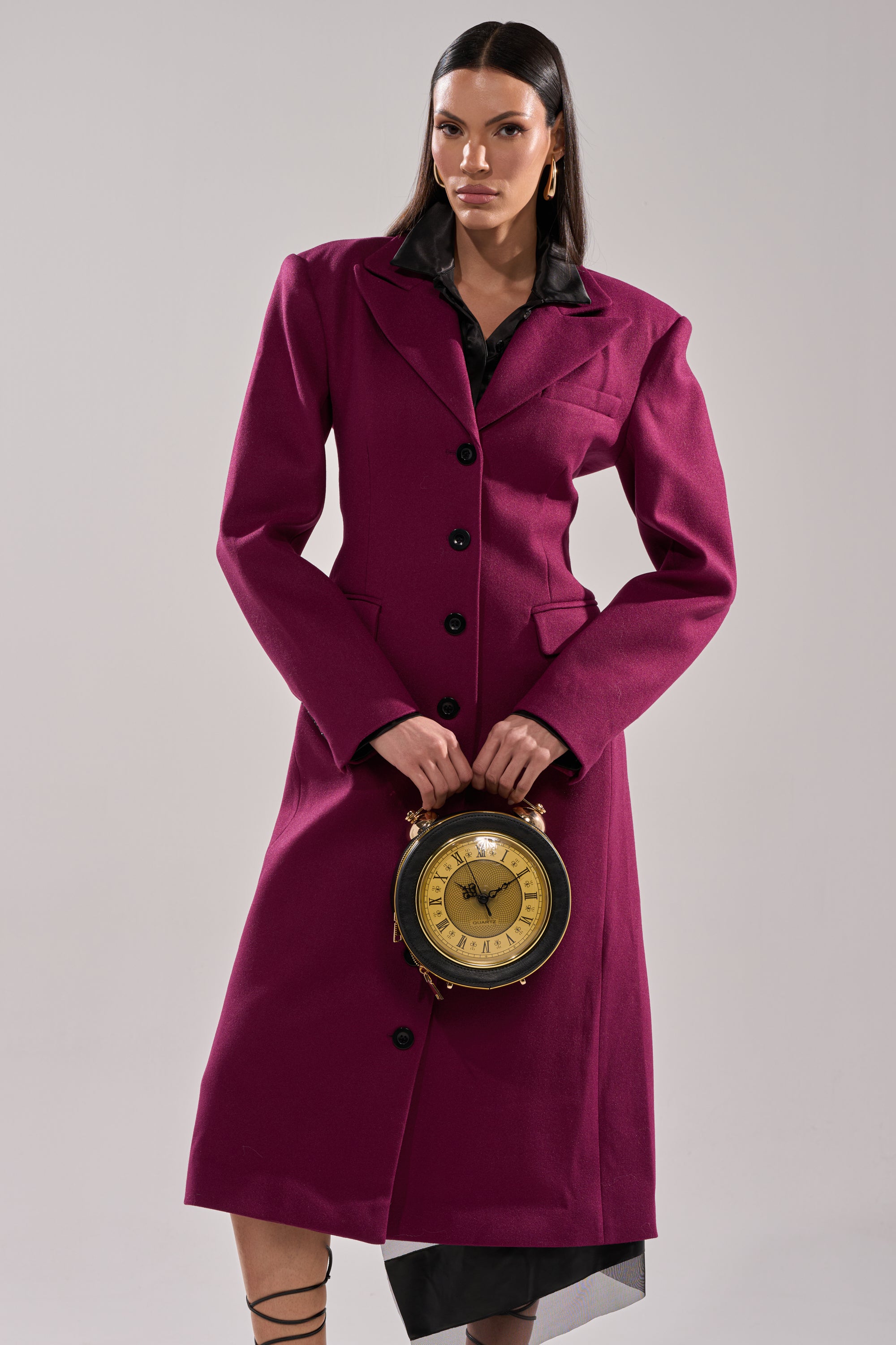 A woman in a long SCARLETT LUXE TRENCH IN PURPLE stands against a plain background, holding a round vintage clock with both hands. She has straight dark hair and wears strappy black shoes.