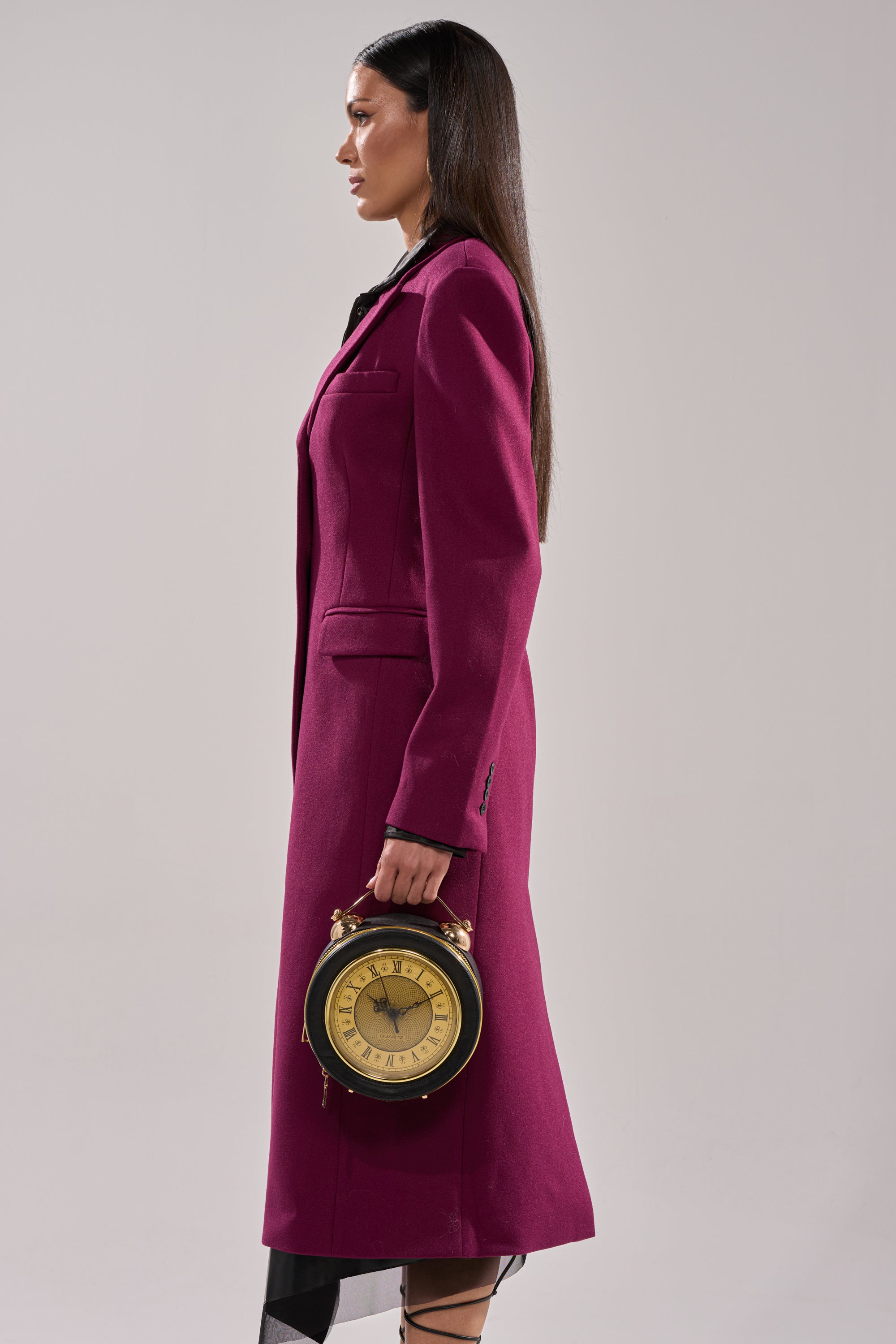 A woman in the SCARLETT LUXE TRENCH IN PURPLE stands in profile against a plain background, holding a round, vintage-style clock as a handbag by her side.