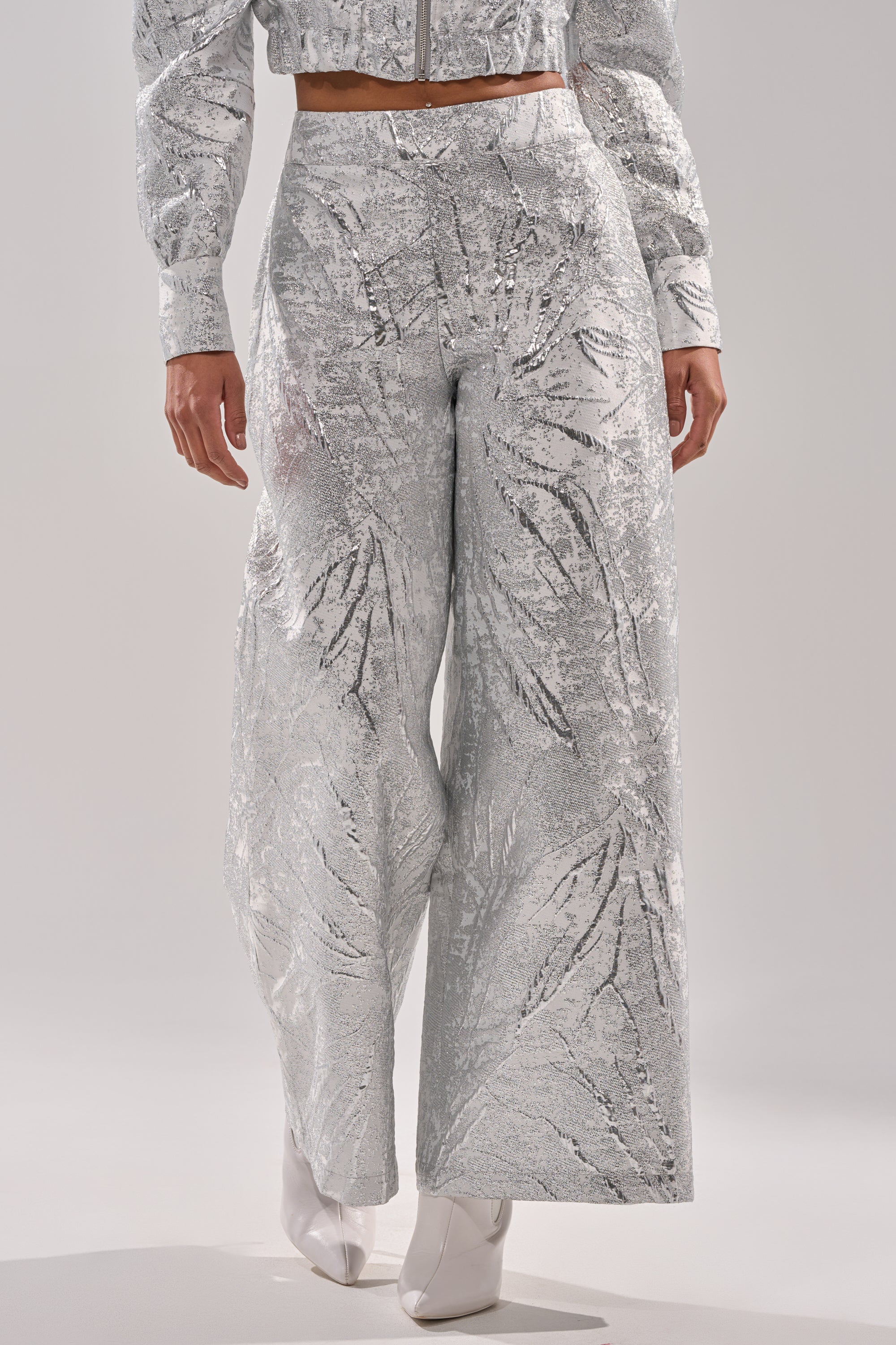 A person wearing the JUST LIKE THAT BROCADE PANT—high-rise, wide-leg pants in silver metallic with a leaf-like pattern—paired with a matching cropped jacket and white pointed-toe shoes. 0