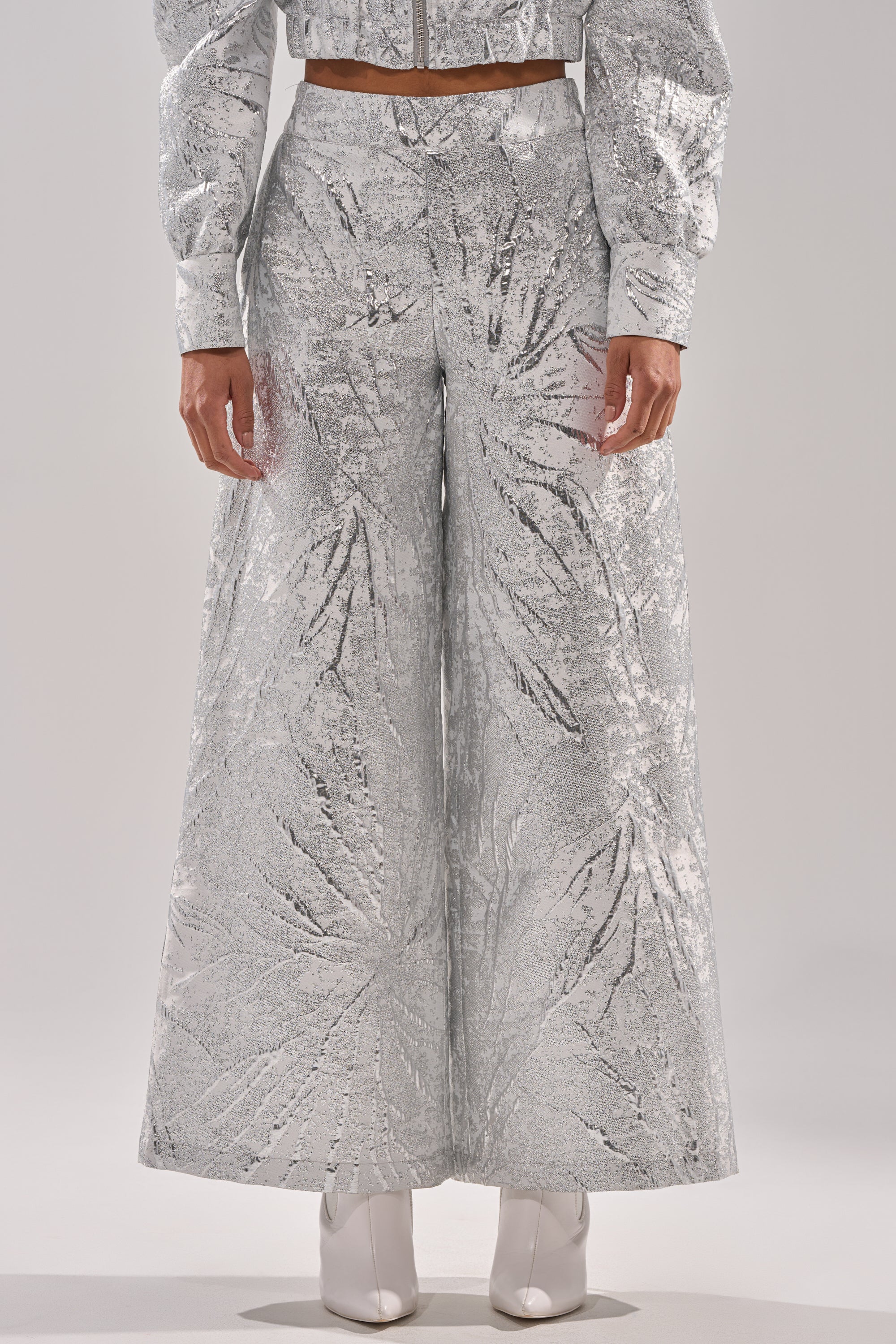 Wearing the JUST LIKE THAT BROCADE PANT in a silver wide-leg silhouette with a leaf pattern, plus matching jacket sleeves and white pointed-toe boots, a person stands against a plain light background. 1
