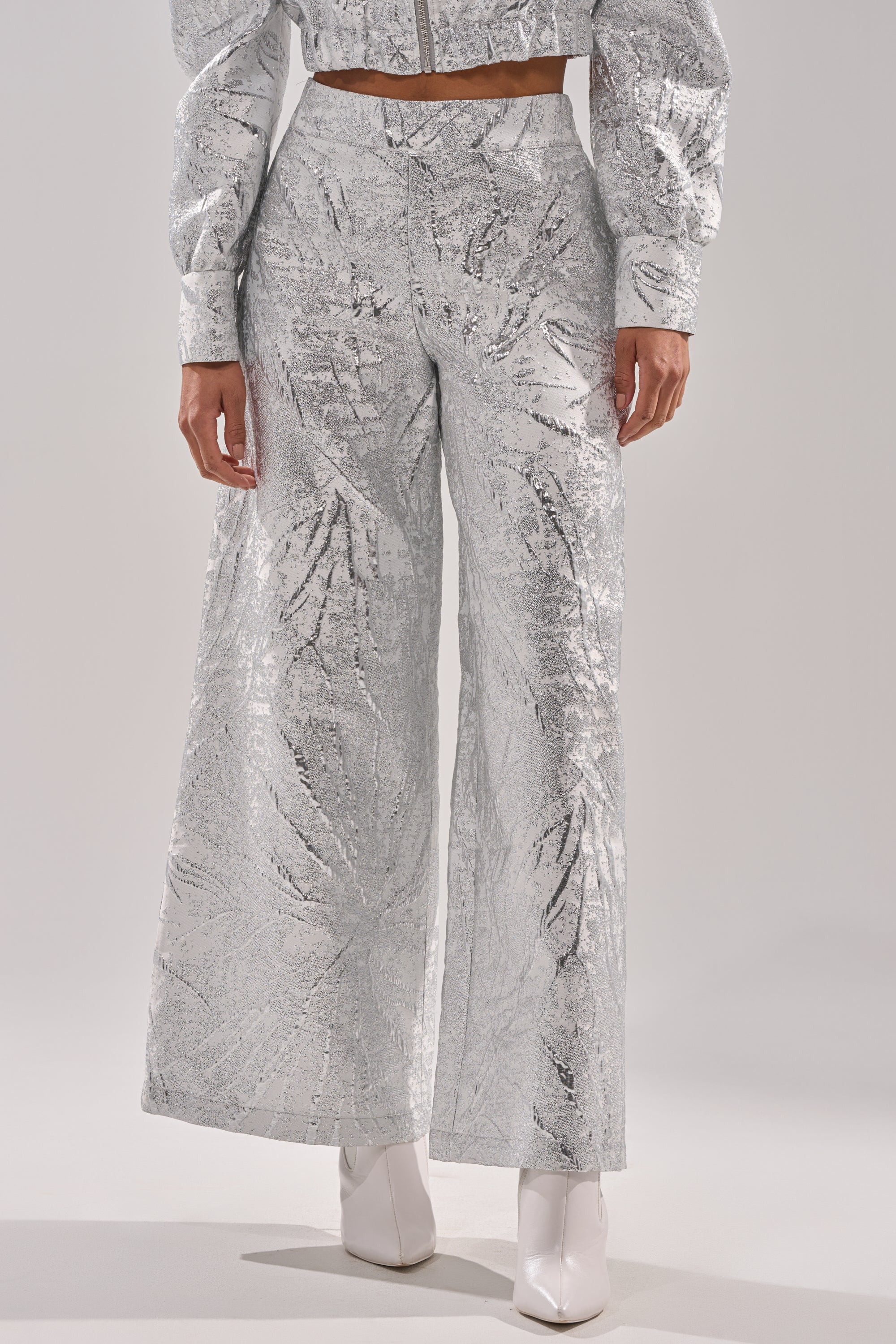 Wearing the JUST LIKE THAT BROCADE PANT, a person stands against a plain light background, styled with wide-leg silver sequins, a matching cropped jacket, and white ankle boots. 2