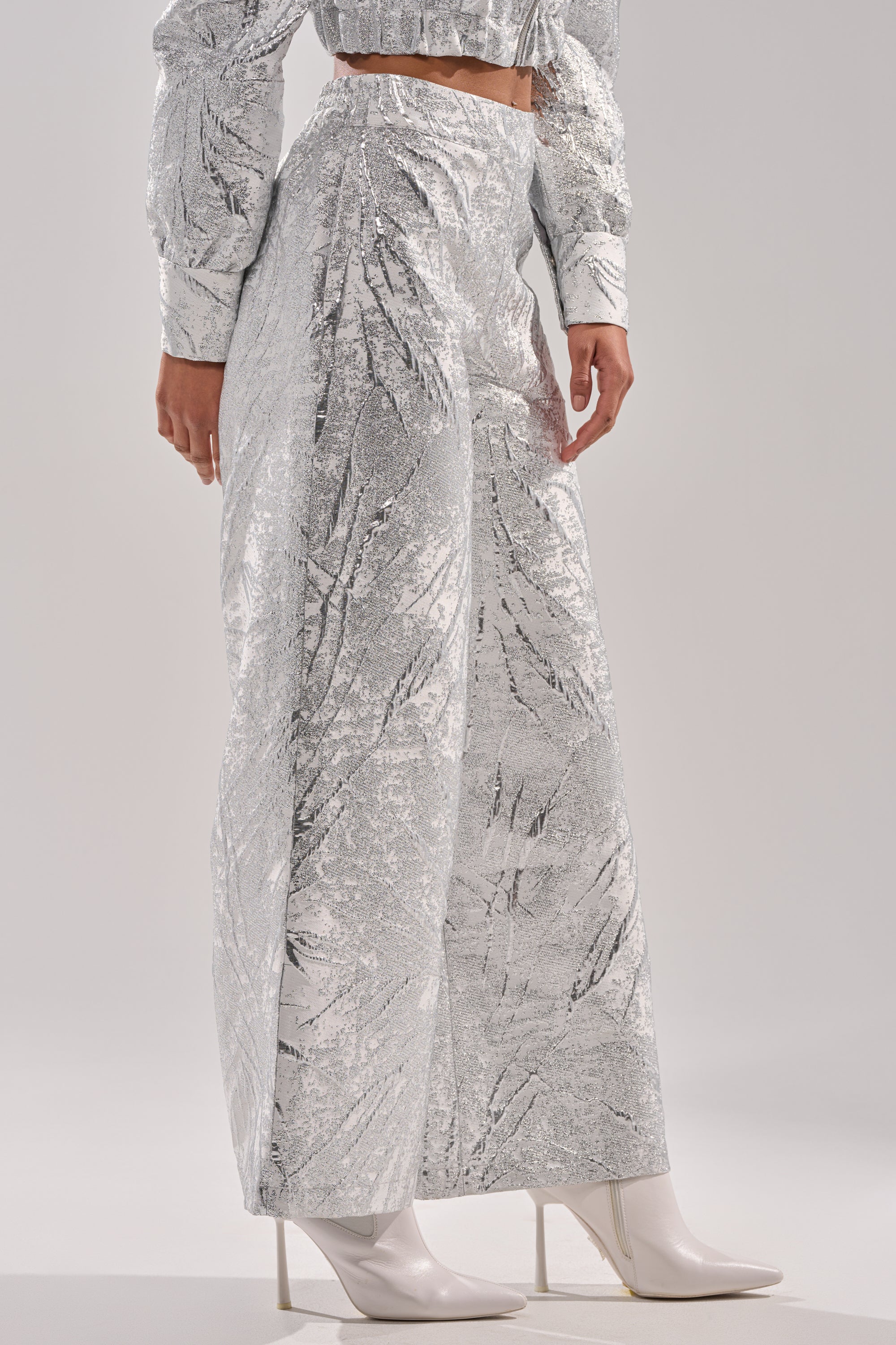 Someone wears the JUST LIKE THAT BROCADE PANT—high-rise, wide-leg with a shiny metallic silver texture—paired with a matching cropped top and white pointed-toe heeled boots for a striking, reflective look. 3