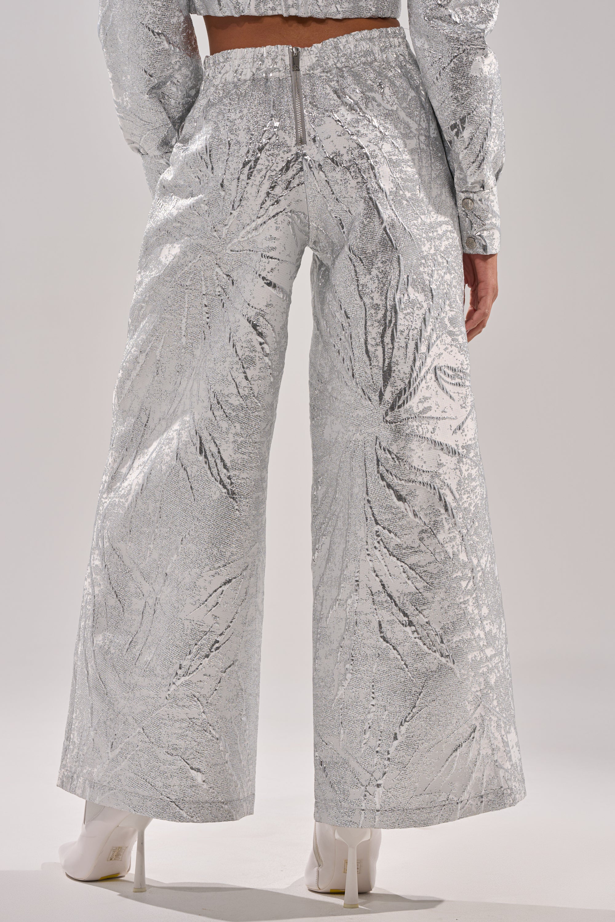 A person is shown from the back wearing the JUST LIKE THAT BROCADE PANT—high-rise, silver, wide-leg pants with an embossed pattern, visible waist zipper, and shiny texture—paired with white pointed-toe shoes. 4
