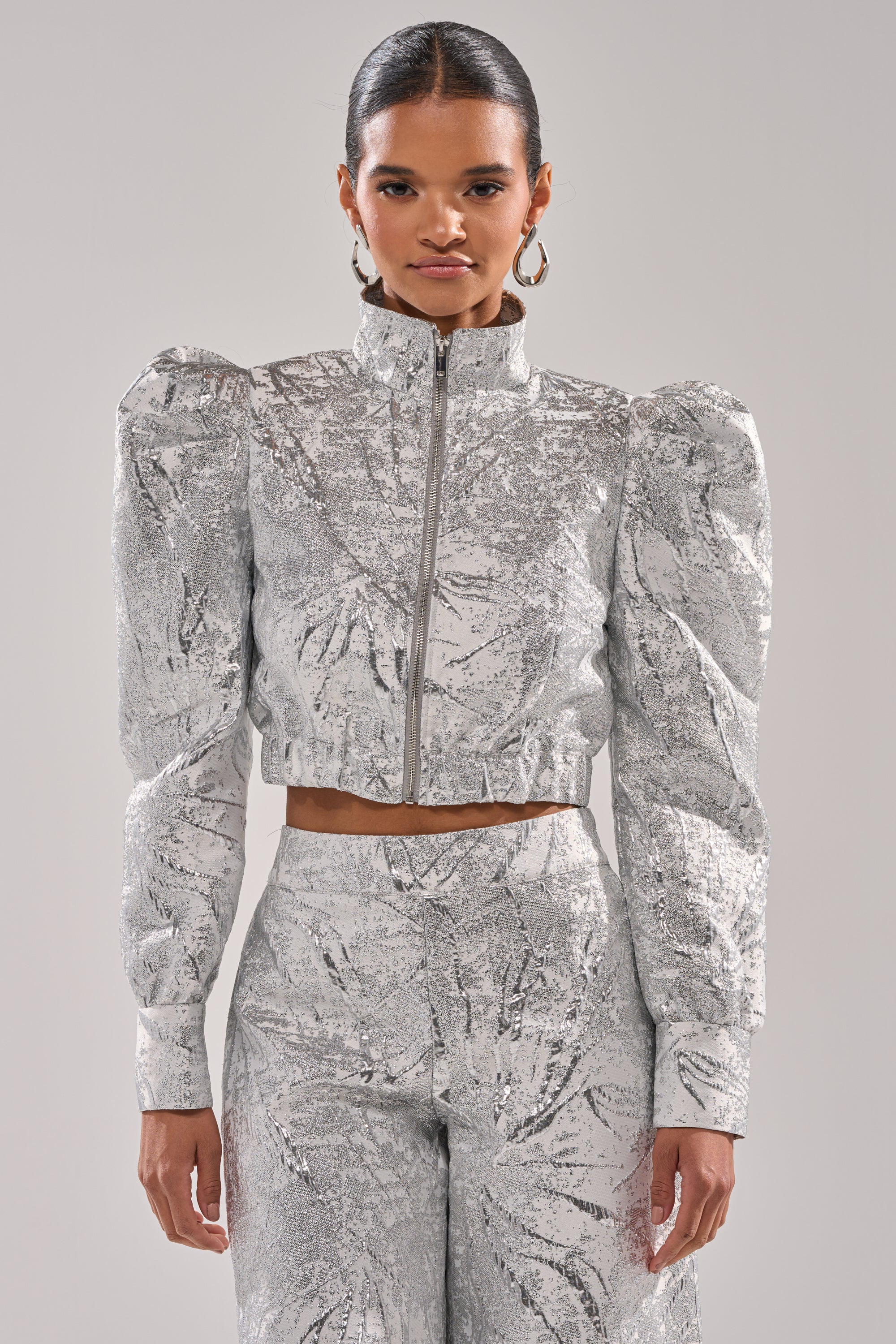 A woman wears the JUST LIKE THAT BROCADE BOMBER with puffed shoulders and matching high-waisted pants, standing against a plain light gray backdrop. Her hair is pulled back, and she accessorizes with hoop earrings. 1