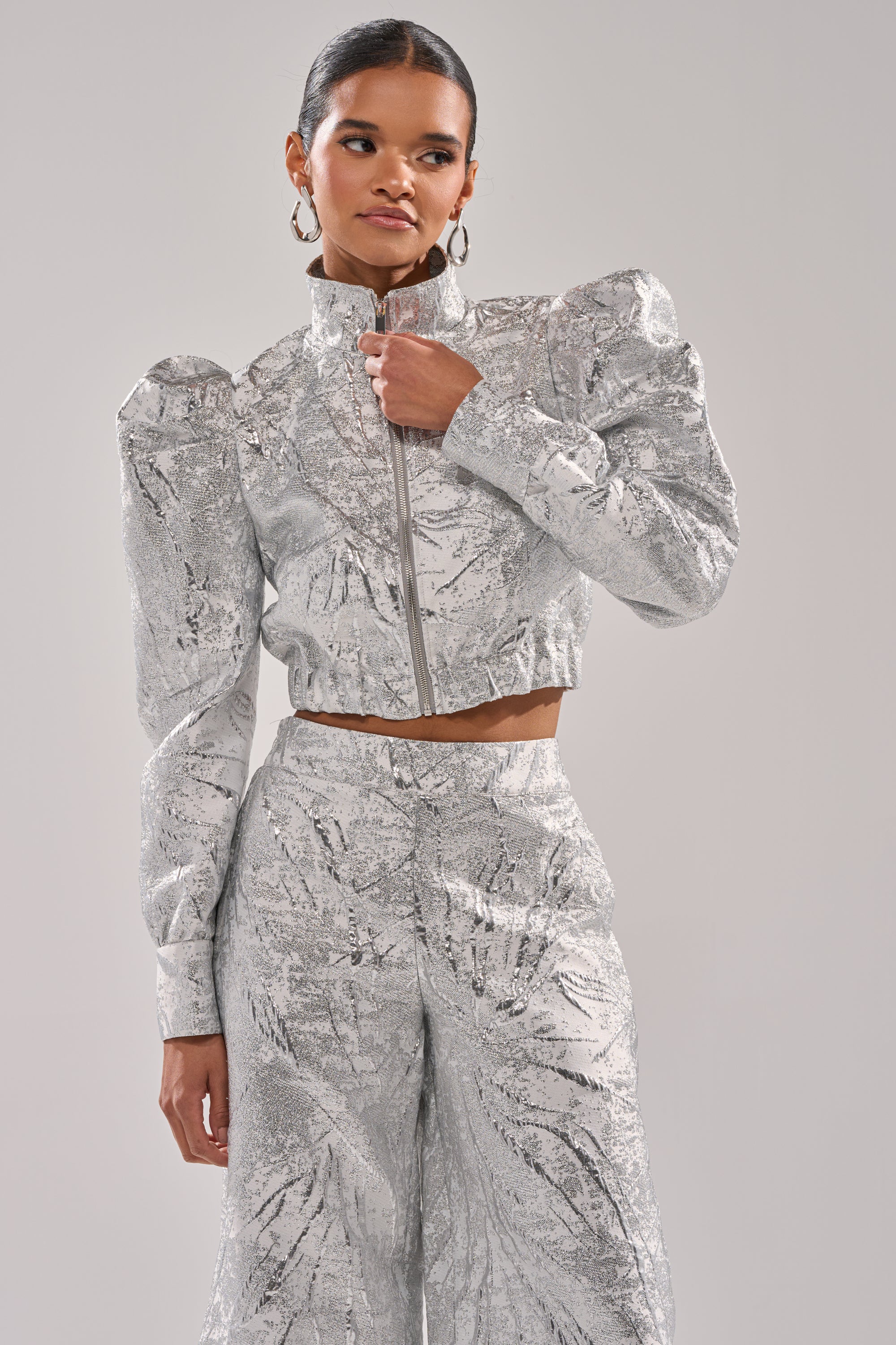 A woman wears the JUST LIKE THAT BROCADE BOMBER—a shiny, silver metallic cropped jacket with puffed shoulders—paired with matching high-waisted pants, standing against a plain light gray background. 2