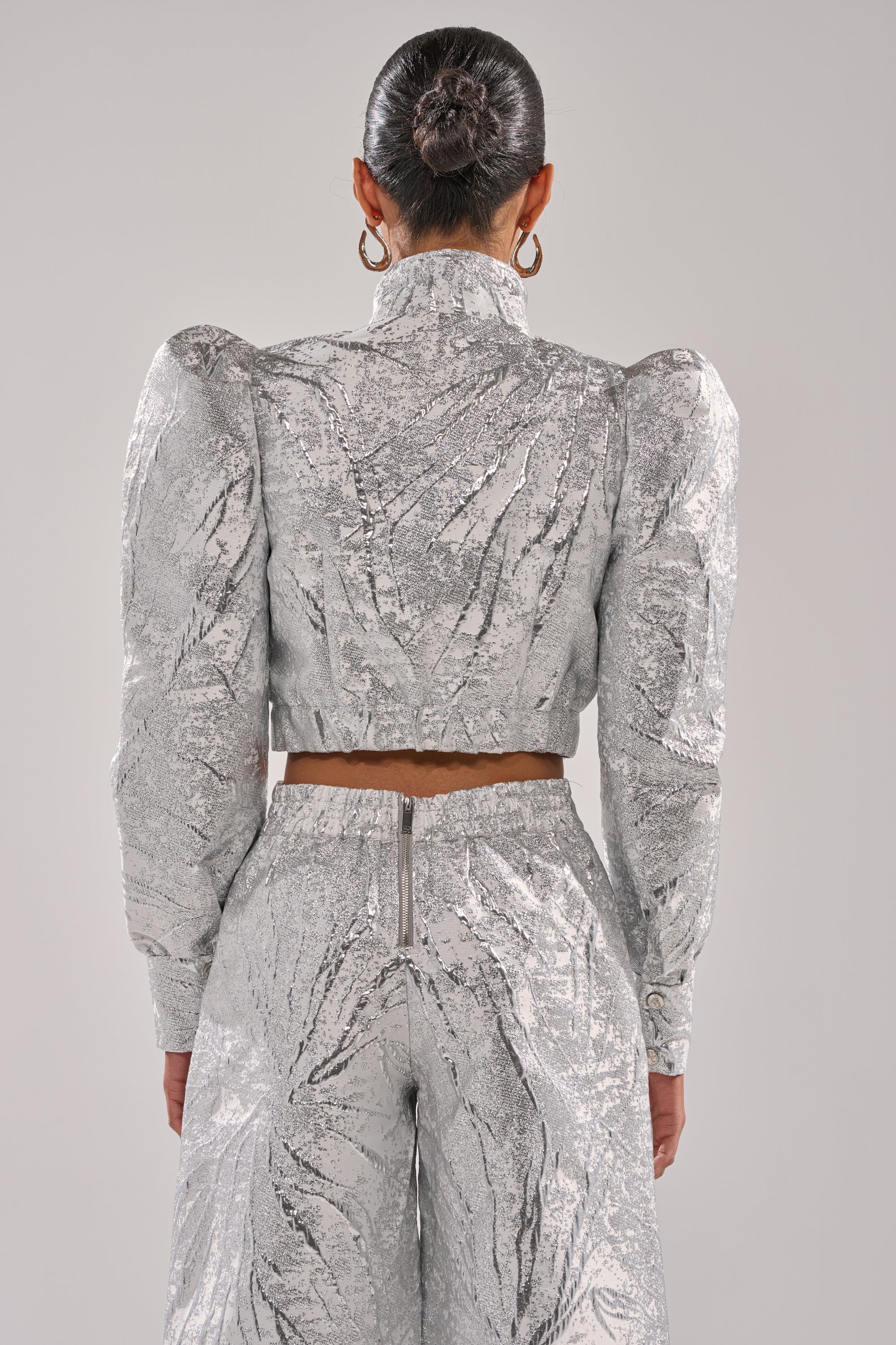 A person with hair in a sleek bun, shown from the back against a plain light background, wears the JUST LIKE THAT BROCADE BOMBER featuring shiny metallic fabric and puffed shoulders paired with matching high-waisted pants. 4