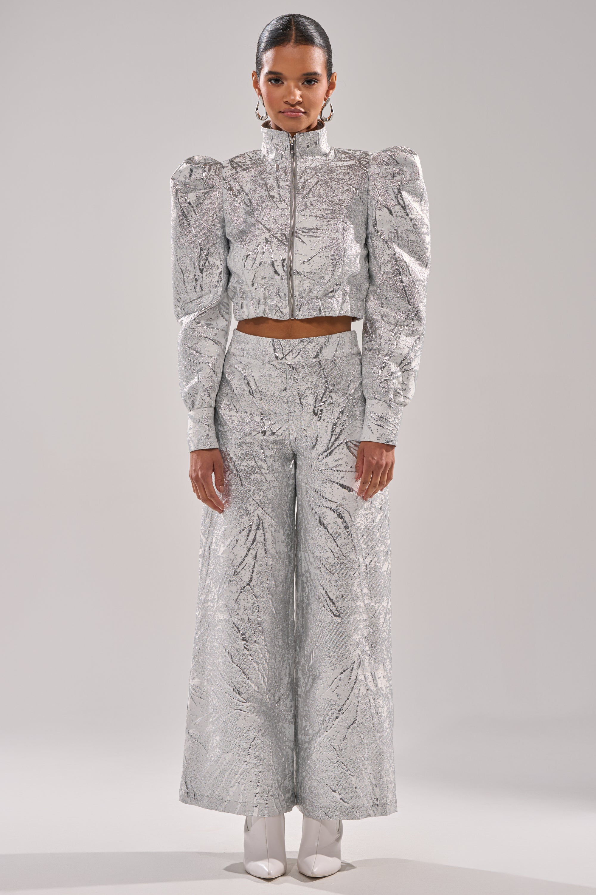 A woman faces forward in the JUST LIKE THAT BROCADE BOMBER, a metallic cropped jacket with puffed shoulders, paired with matching high-waisted wide-leg pants and white pointed-toe boots against a plain light gray background. 5