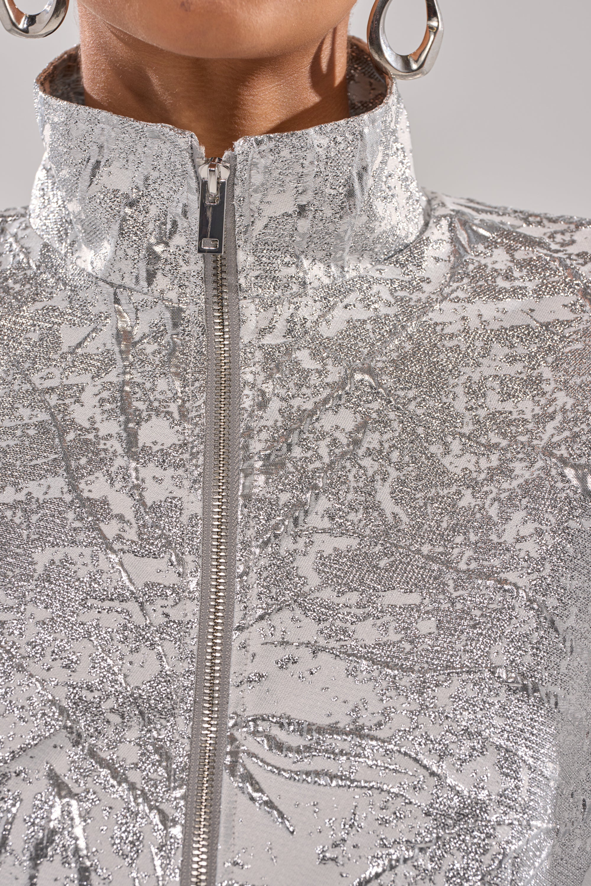 Close-up of a person wearing the JUST LIKE THAT BROCADE BOMBER, featuring a high collar, metallic brocade fabric with zipper and shimmering glitter texture, paired with large round silver hoop earrings. 7