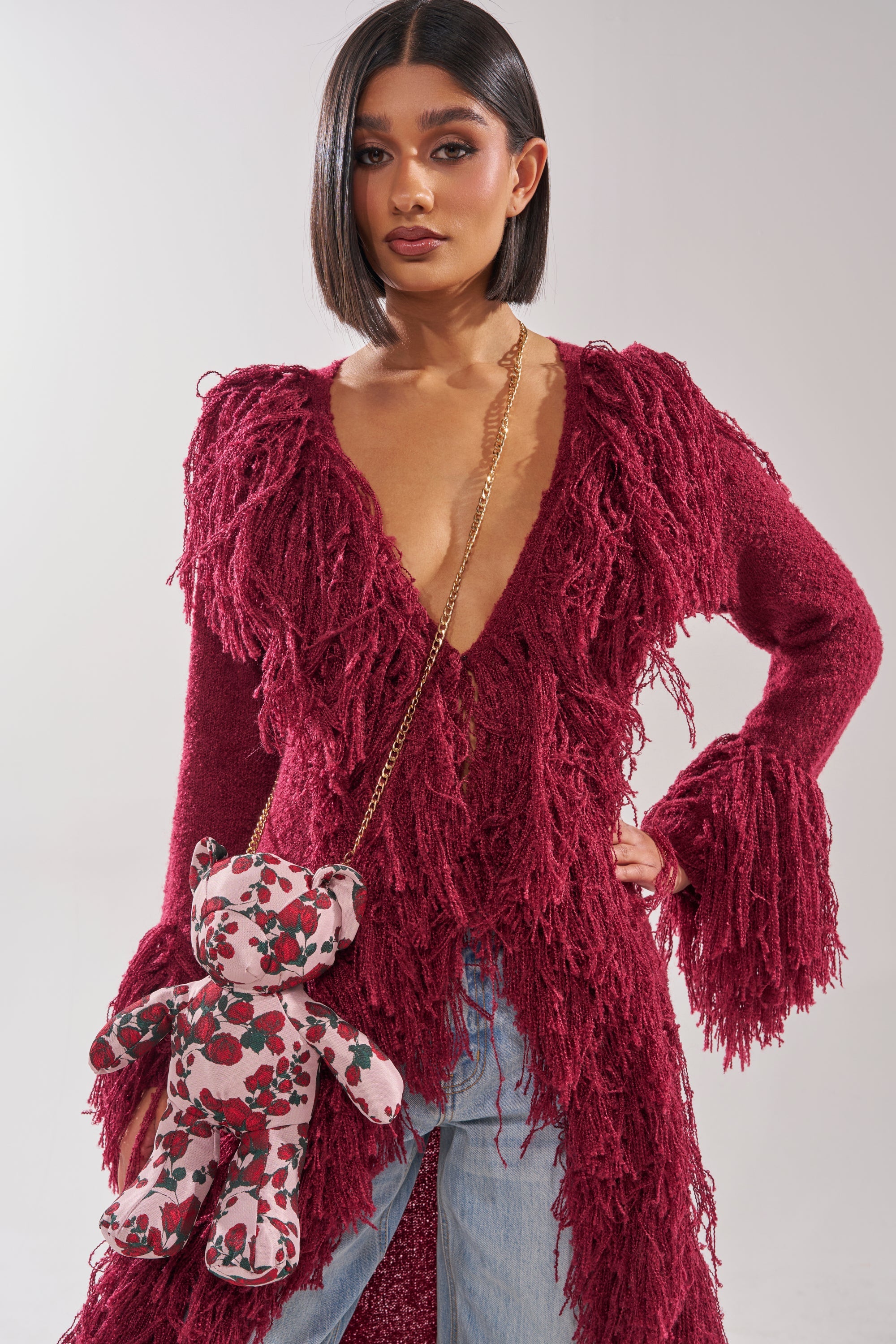 A woman with a sleek bob wears a deep red, fringed V-neck sweater and light blue jeans. She carries THE ONE ROSE BEAR BAG—a teddy crossbody with a gold chain strap—over her shoulder, standing against a plain background. 1