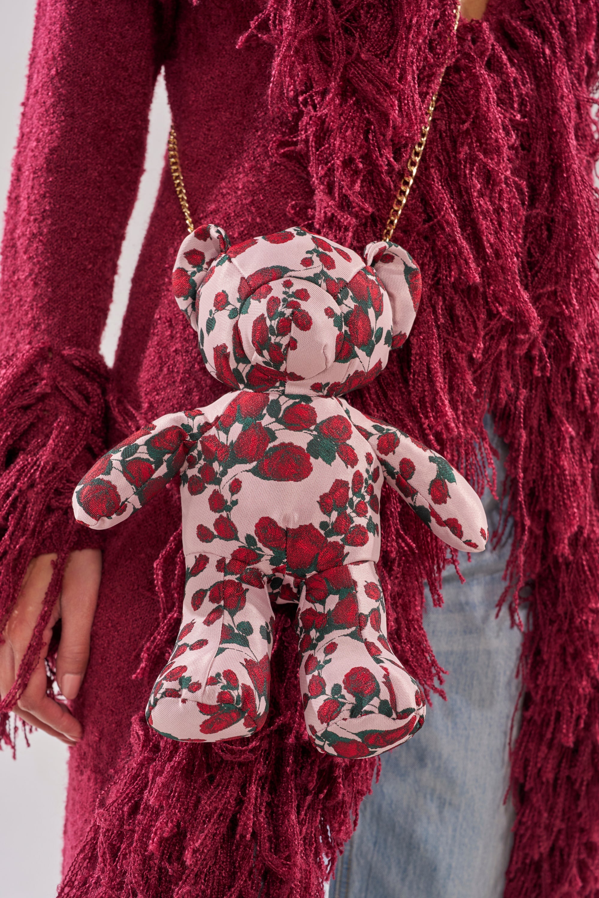 A person in a textured, fringed burgundy cardigan and blue jeans carries THE ONE ROSE BEAR BAG—a small teddy crossbody with a pink and red floral pattern and gold chain strap. 5