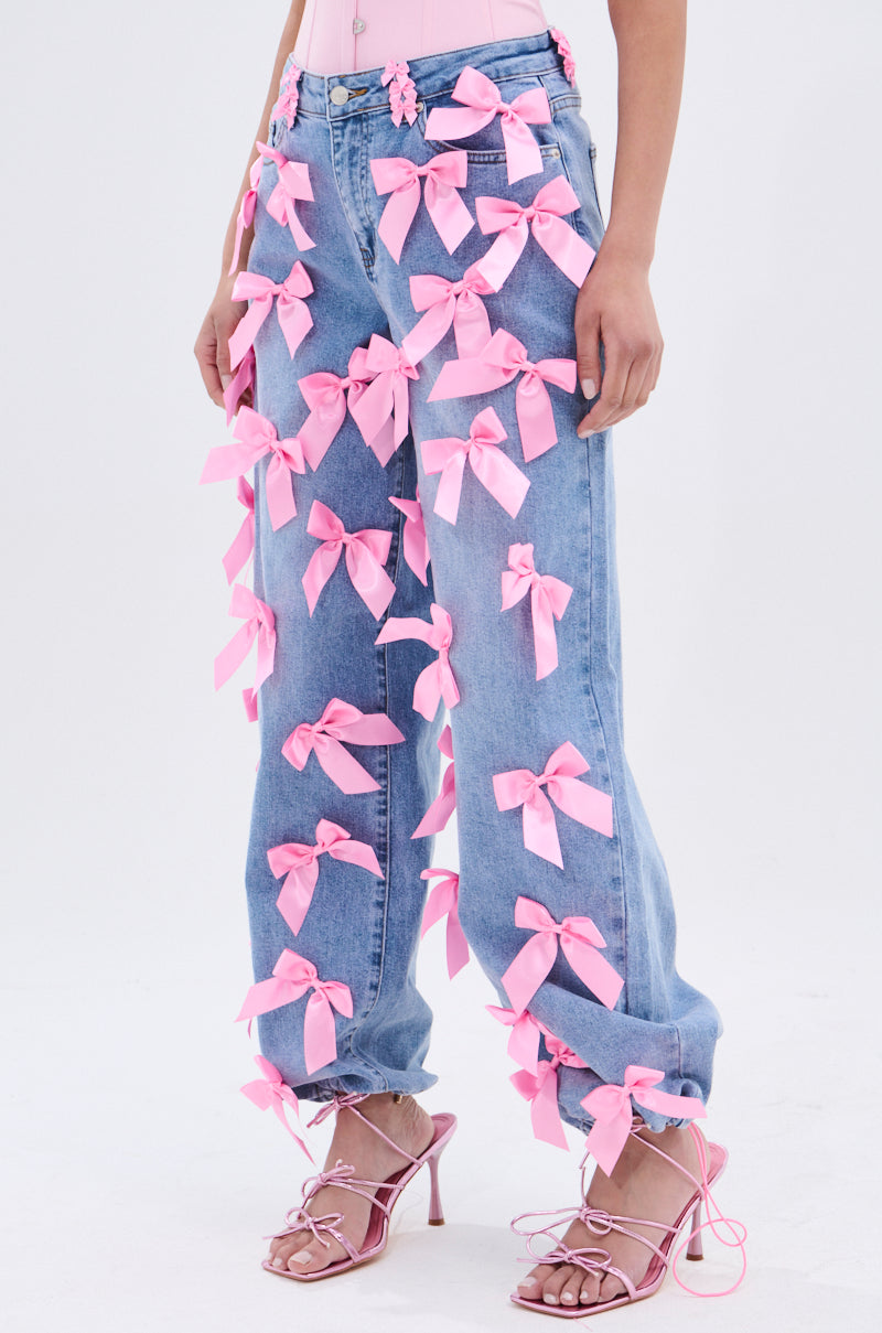 Someone wears PRETTY IN PINK BOW DETAIL DENIM JEANS, styled with pink strappy high-heeled sandals and a matching pink top, against a plain light-toned background.