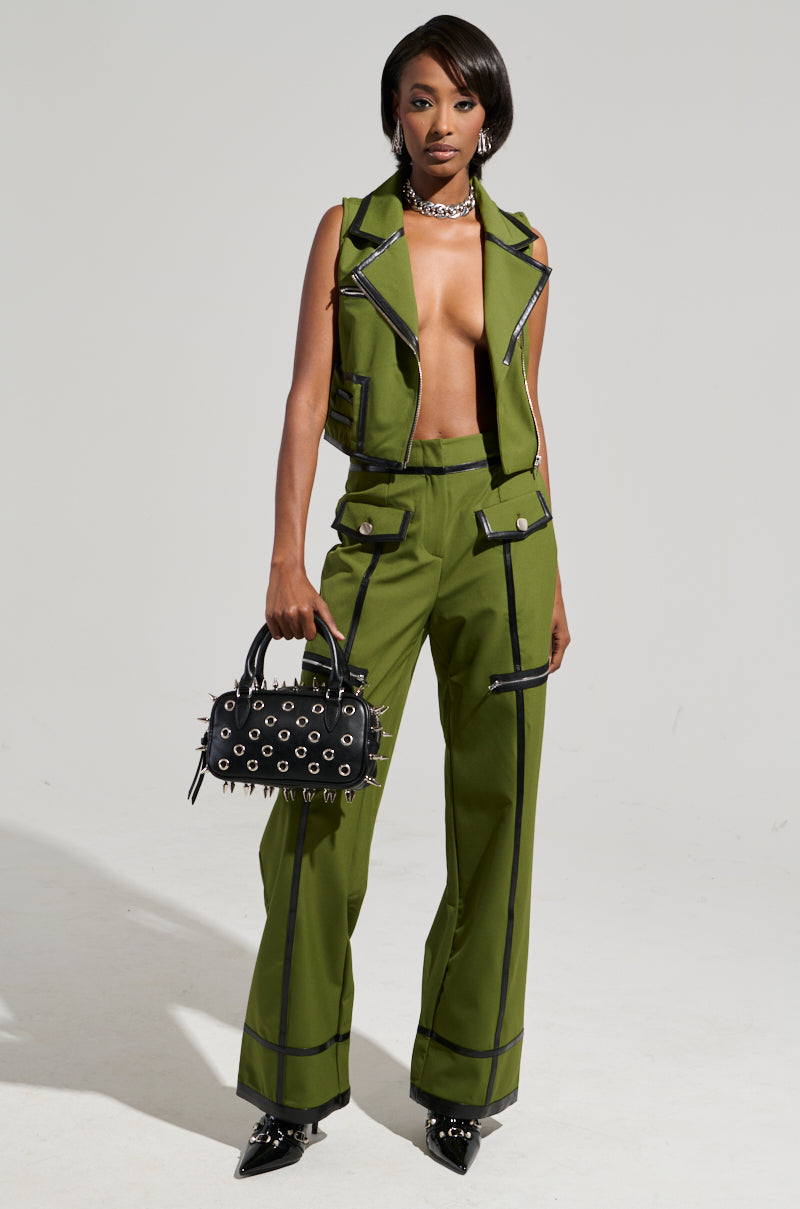 Wearing the EAST HAMPTON WIDE LEG TROUSERS paired with a coordinating green sleeveless jacket featuring black faux leather trim, an individual exudes confidence. They carry a stylish black handbag embellished with eyelet details and complete the look with sleek black pointed shoes.