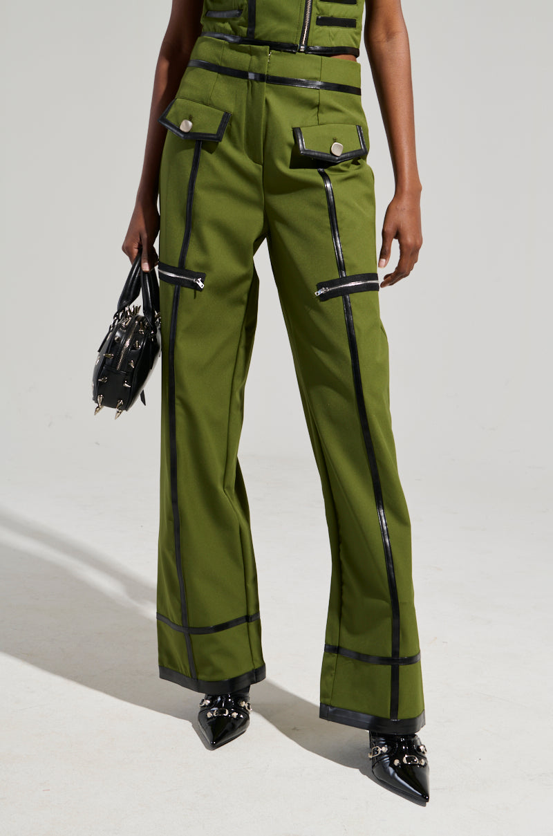 A person wearing EAST HAMPTON WIDE LEG TROUSERS in green, featuring black accents and zipper details, stands holding a black handbag. They are also wearing pointed black shoes adorned with silver embellishments. The background is plain and light-colored.