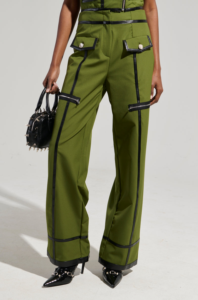 A person dressed in olive green EAST HAMPTON WIDE LEG TROUSERS, featuring black accents and large buttons, is carrying a small black handbag adorned with silver studs. The trousers have an edgy touch with their faux leather trim. They complete the look with pointed black shoes.