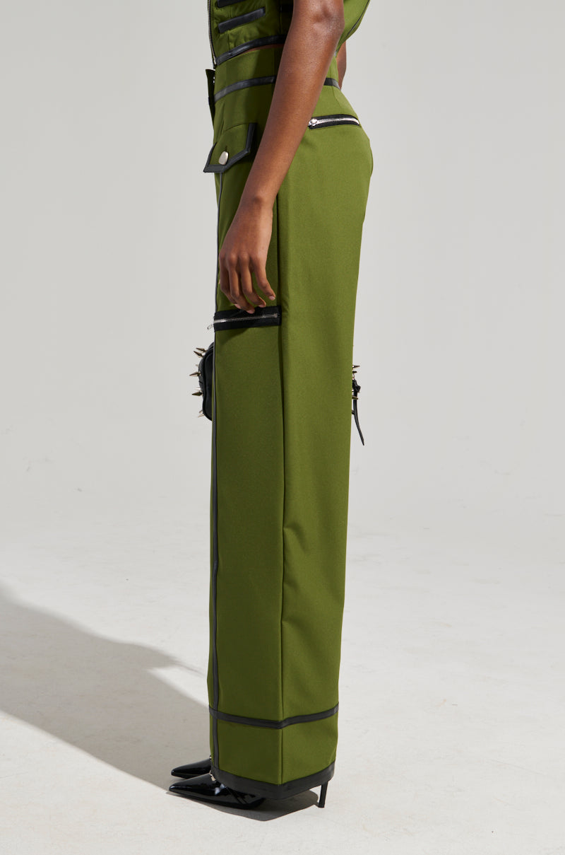 A model is displayed from the side wearing the EAST HAMPTON WIDE LEG TROUSERS in olive green, characterized by black accents and silver studs. These high-rise pants are detailed with rectangular embellishments, faux leather trim, and multiple zippers, set against a plain light gray background.