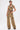 A woman poses against a plain white background wearing the EAST HAMPTON WIDE LEG TROUSERS IN BROWN, featuring a mid-rise fit, wide legs, faux leather piping, and large pockets.