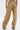 A person wears the EAST HAMPTON WIDE LEG TROUSERS IN BROWN, featuring black faux leather piping on the back pockets, standing against a plain white background.