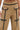 Close-up of a person wearing the EAST HAMPTON WIDE LEG TROUSERS IN BROWN, featuring high waist, black faux leather piping, gold buttons and zippers, unique pocket designs, and a partially visible tattooed midriff.