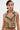 A woman with short, dark hair and star-shaped earrings wears the EAST HAMPTON VEST IN BROWN, showing its asymmetrical front zipper and faux leather piping. Tattoos are visible on her arms and torso as she looks slightly to the side.