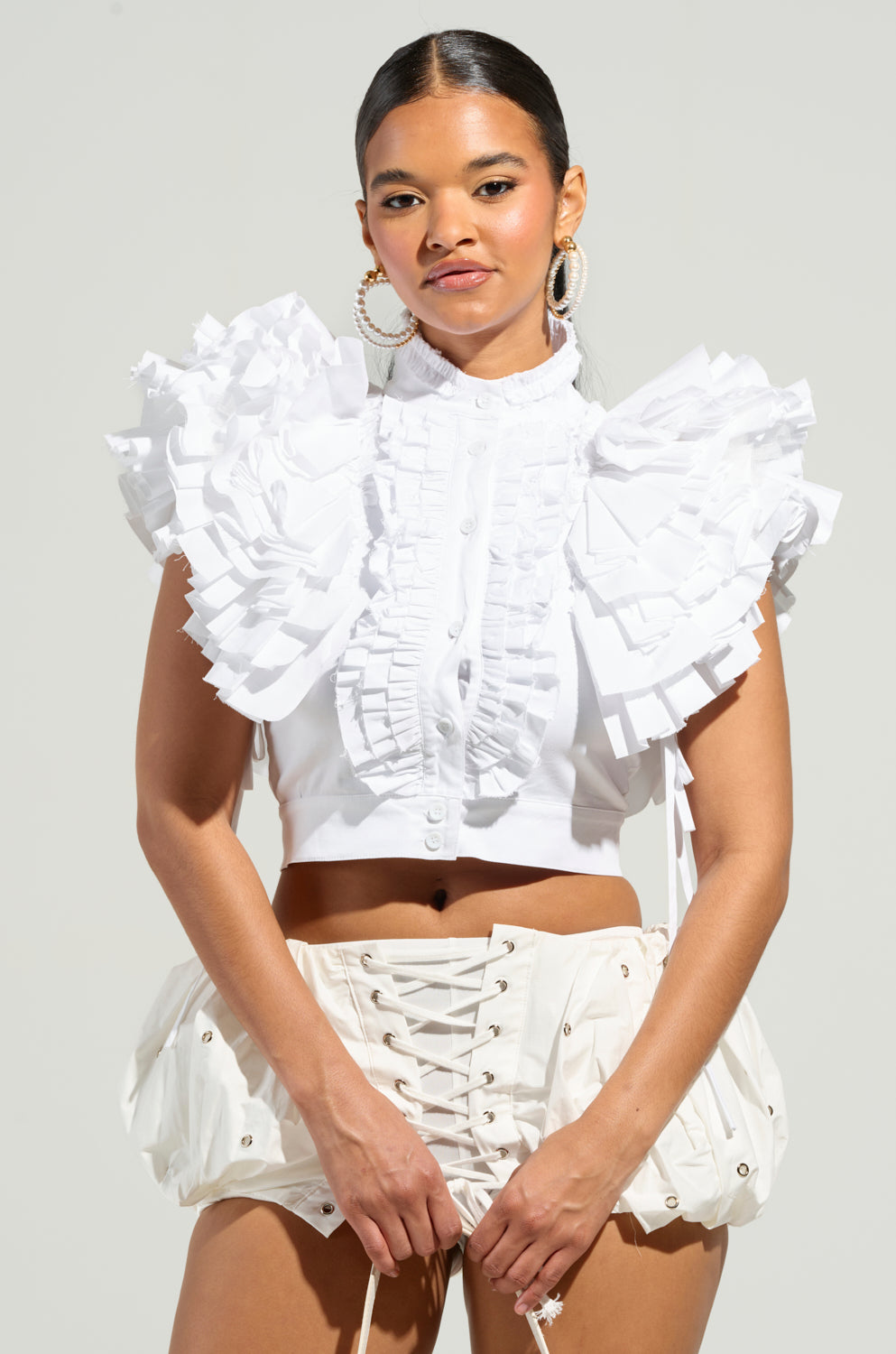 A woman poses confidently in a fashionable ensemble featuring the OVER AND OVER RUFFLE TOP paired with a lace-up white skirt. She accessorizes with hoop earrings, and her long hair is pulled back. The neutral background accentuates her stylish look.