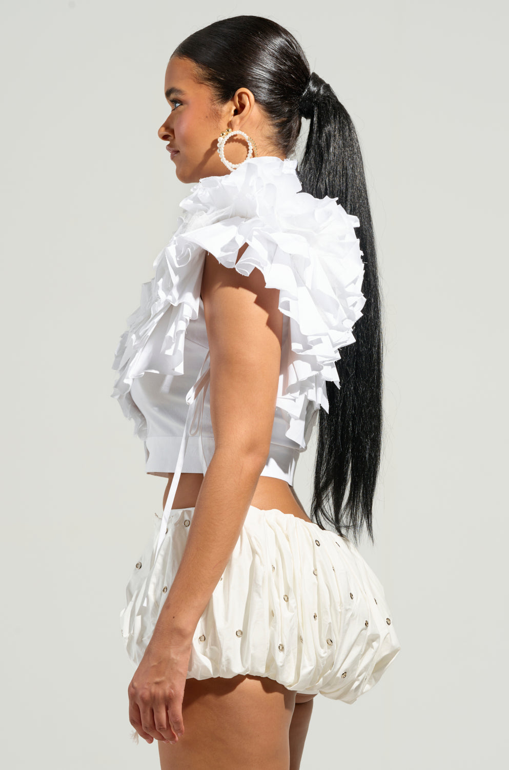 A woman with long black hair is wearing the OVER AND OVER RUFFLE TOP and white bubble shorts. Her outfit, accentuated by the ruffle top, is complemented by large hoop earrings as she stands sideways against a plain background.