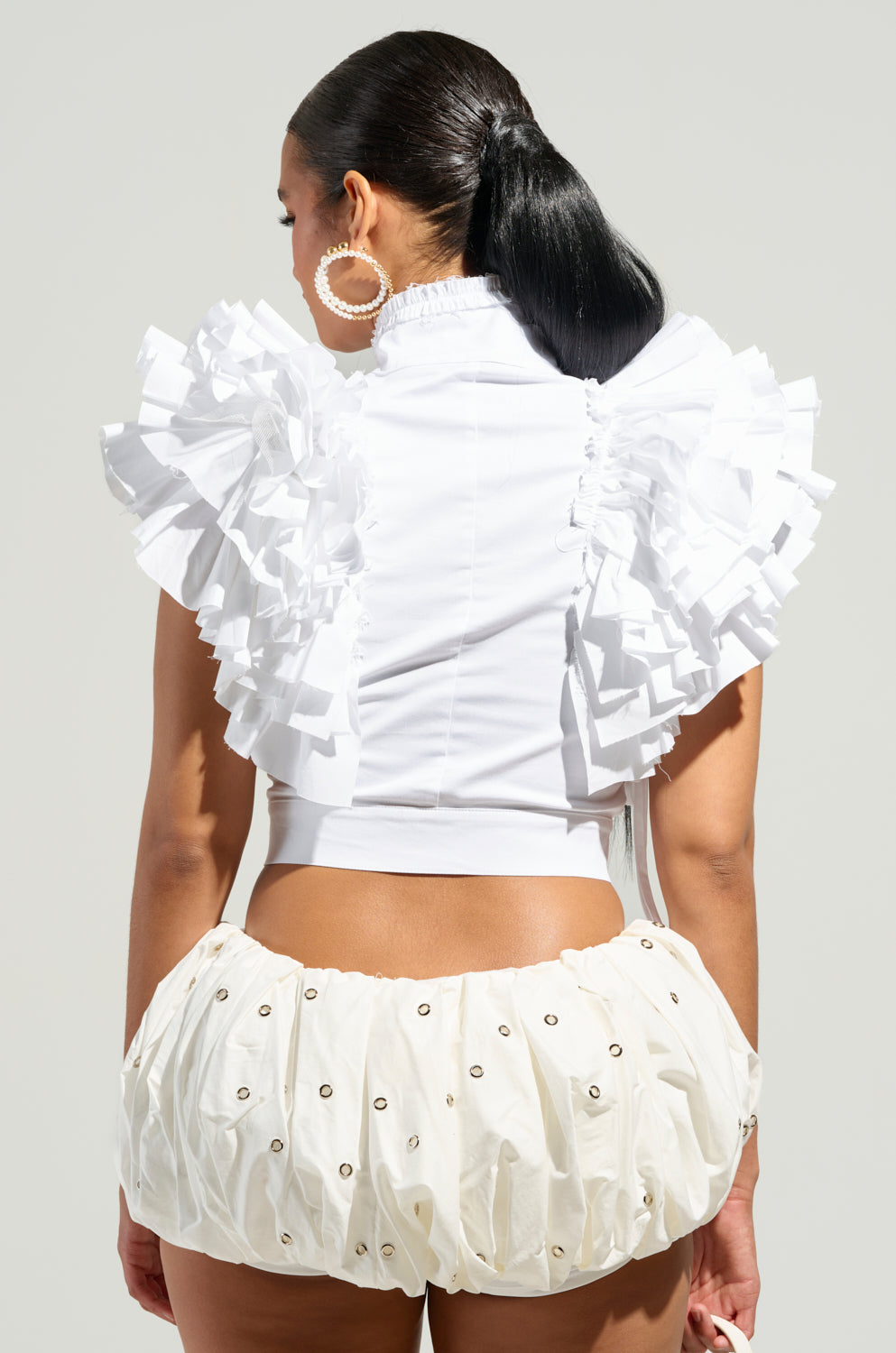 A person with long dark hair is wearing the OVER AND OVER RUFFLE TOP, featuring large ruffled shoulders, paired with a white, embellished bubble skirt that creates a cropped silhouette. They stand against a light gray background.