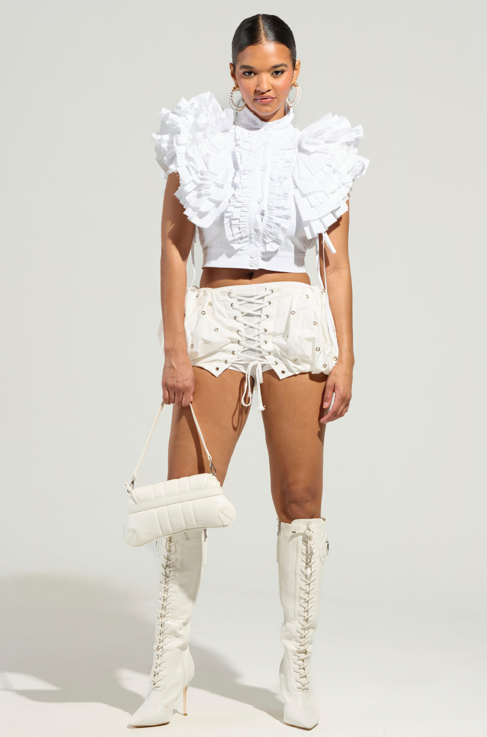 A person is dressed in a chic white ensemble featuring the OVER AND OVER RUFFLE TOP and lace-up shorts, paired with tall lace-up boots. They're holding a stylish white handbag and wearing hoop earrings. The plain grayish background highlights this elegant outfit.