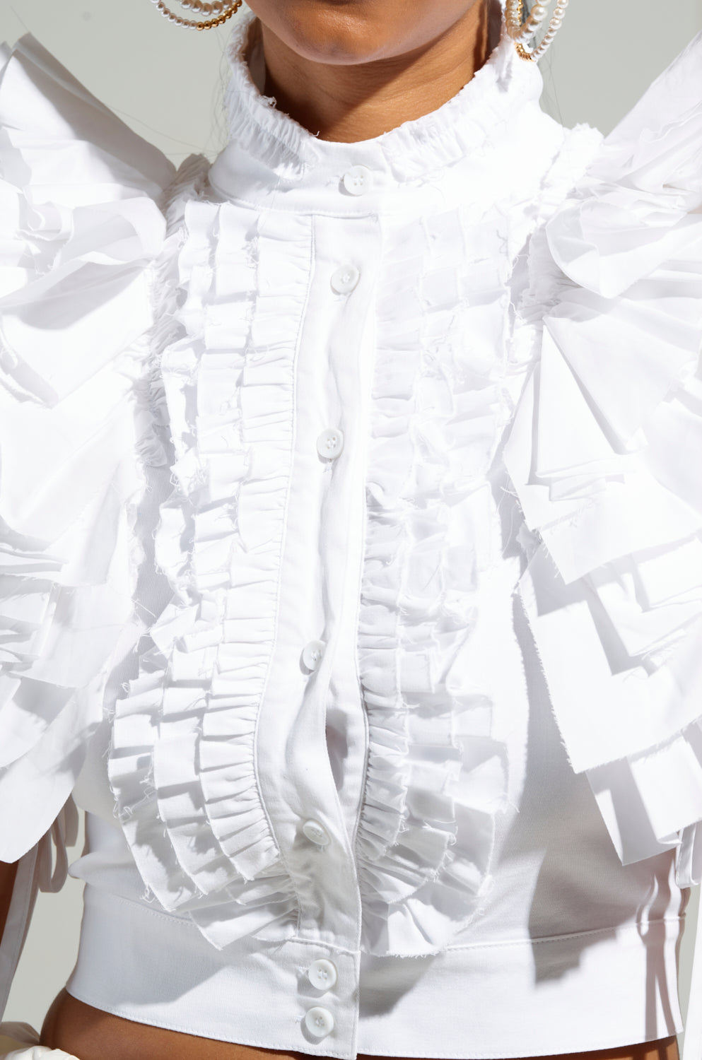 A close-up showcases the OVER AND OVER RUFFLE TOP, featuring an elegant white ruffle design and large, textured sleeves. This button-down beauty pairs perfectly with oversized earrings, while the lighting accentuates the intricate fabric details.