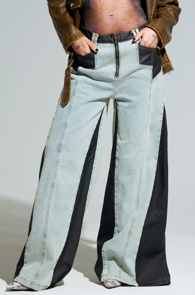 A person is wearing the stylish WILL COLORBLOCK WIDE LEG JEAN, which features chic black side panels and front pockets. This effortlessly fashionable outfit is completed with a brown faux leather jacket, a lace top, and snake-patterned shoes.