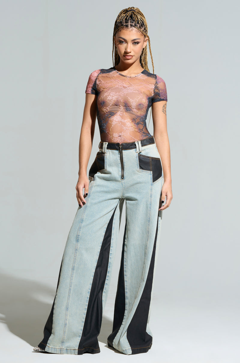 A person with long braided hair is wearing a sheer lace top and the WILL COLORBLOCK WIDE LEG JEAN, featuring high-rise waist and black panels. They stand confidently against a plain background.