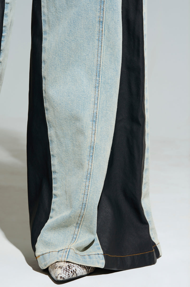 A close-up of a person wearing the WILL COLORBLOCK WIDE LEG JEAN showcases the striking design, with one side in light blue denim and the other in black fabric. The high rise waist enhances its modern appeal, while snake-patterned shoes peek out stylishly from below.