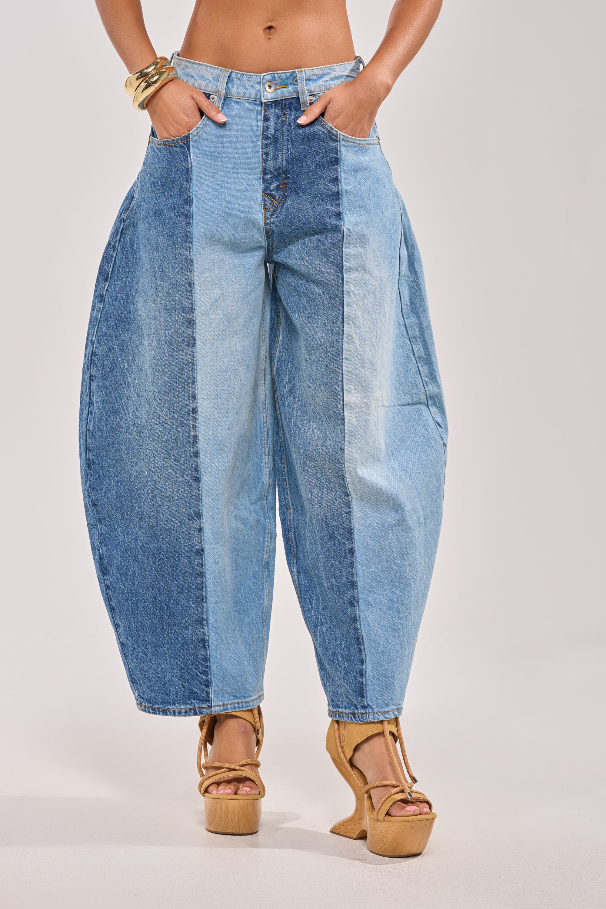 Wearing the MIXED EMOTIONS COLORBLOCKED BARREL DENIM in blue shades, paired with tan platform heels and gold bracelets, a person stands with hands in pockets against a plain backdrop. 0