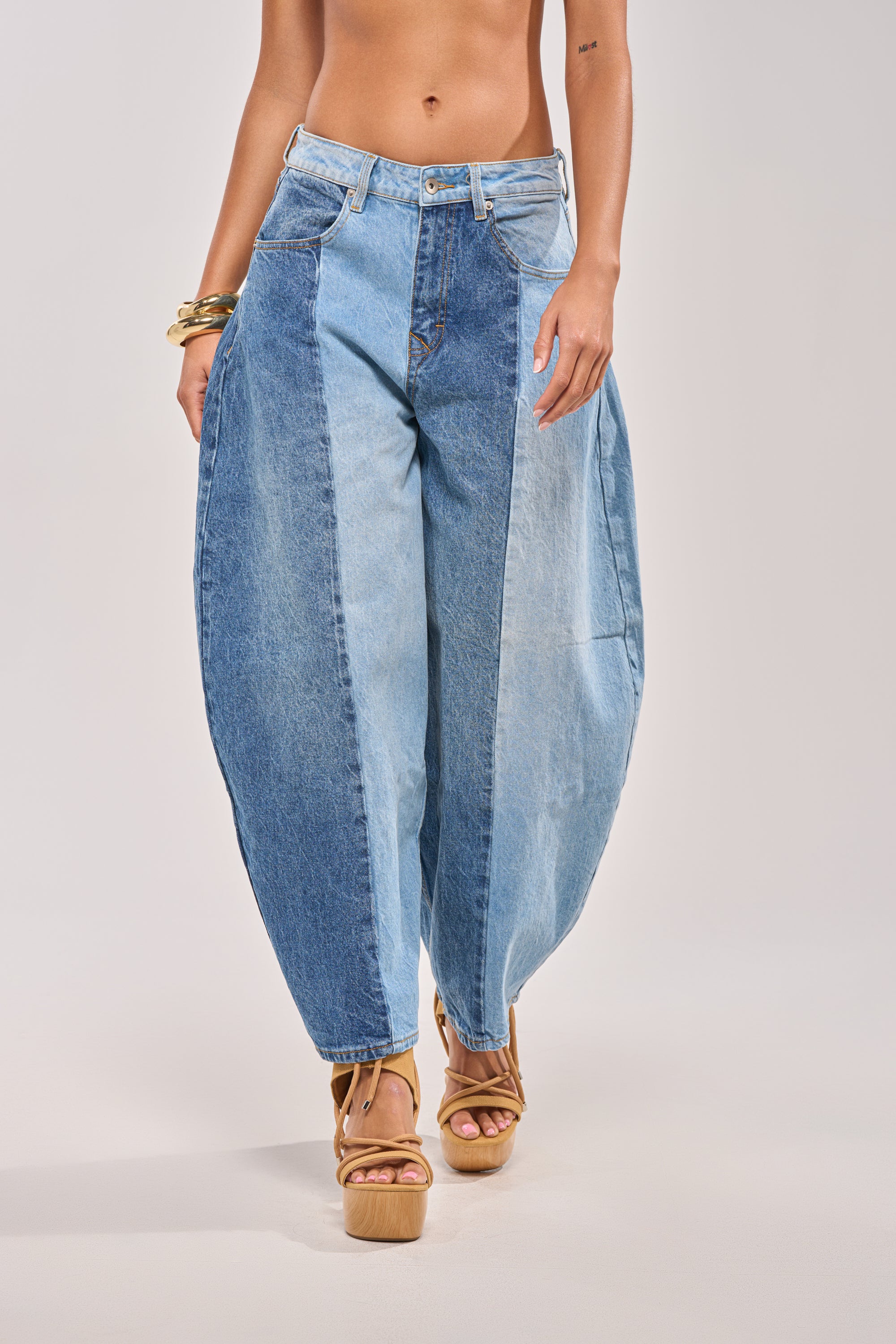 A person shown from mid-torso down wears the MIXED EMOTIONS COLORBLOCKED BARREL DENIM, featuring exaggerated curved sides, styled with tan platform sandals and gold bangles against a plain background. 1