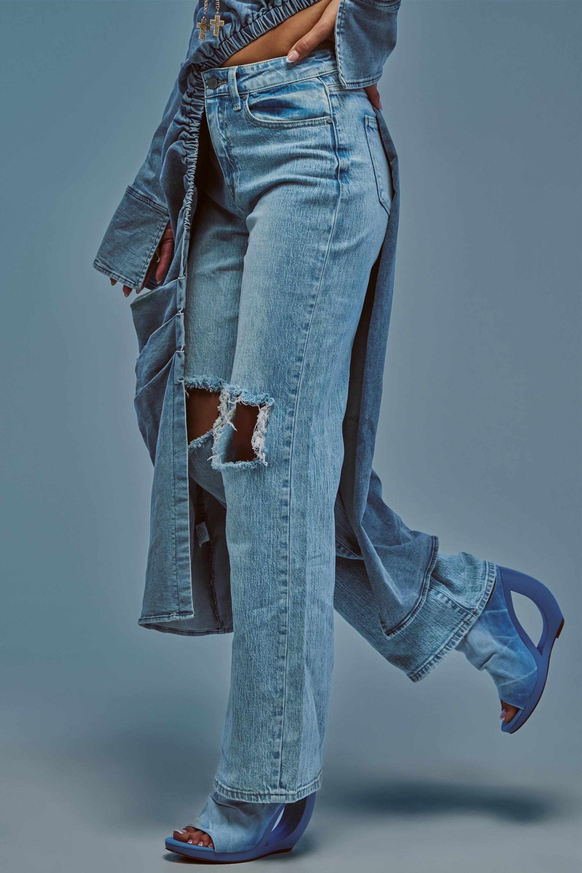 Wearing the SIMPLE AND GOOD RELAXED DENIM, a person poses against a blue background with one leg bent back and a hand resting in the pocket, styled with a denim jacket and blue high-heeled sandals.