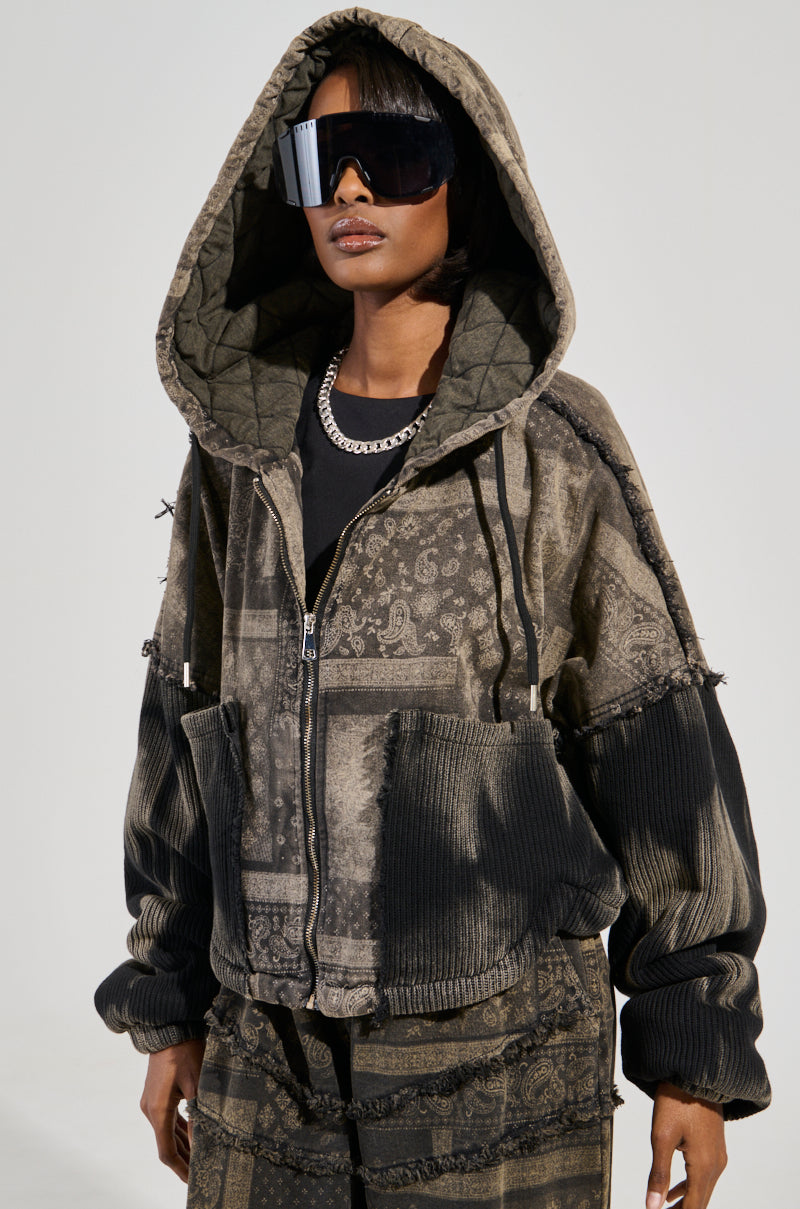 A person wearing a Phoenix Hooded Bomber with a quilted texture stands against a plain background. Their jacket is paired with large sunglasses, a metallic chain necklace, and matching pants.