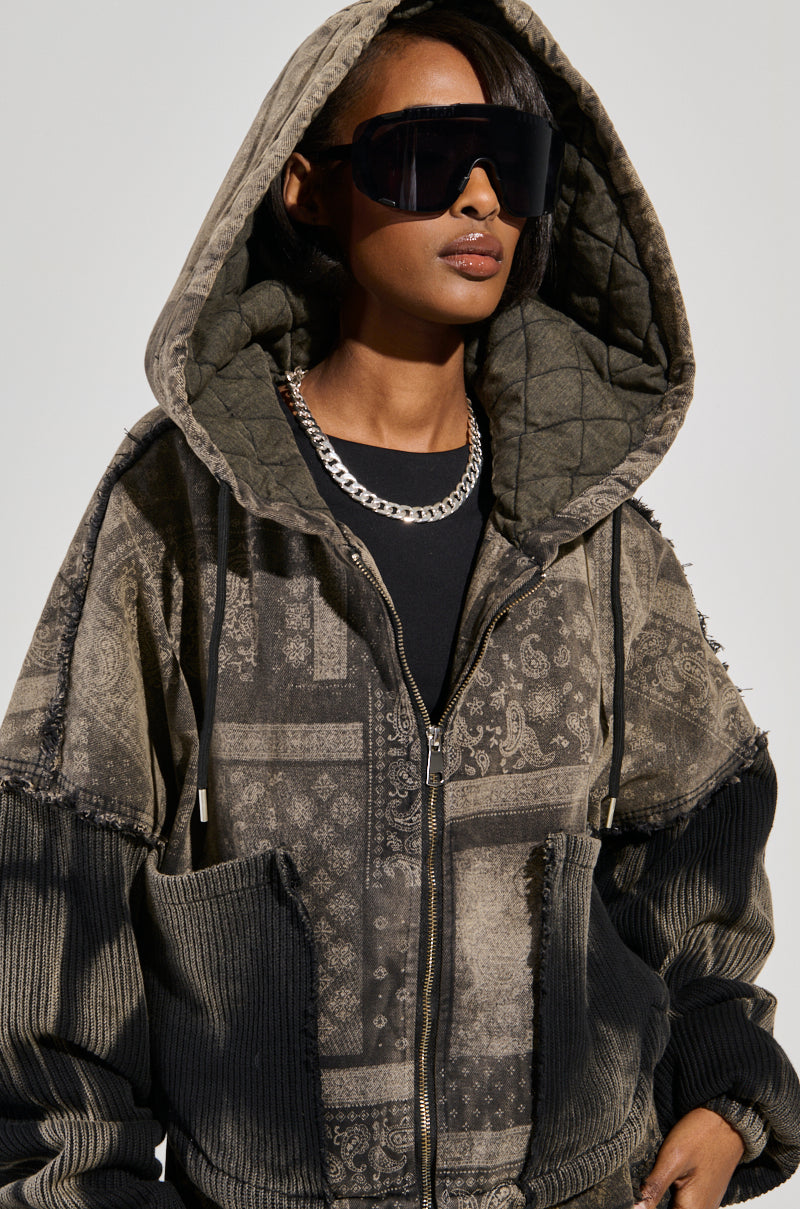 A person wearing a large quilted hoodie with a paisley print design and dark oversized sunglasses. Their silver chain necklace gleams as they look off to the side against a simple background, exuding style in their PHOENIX HOODED BOMBER.