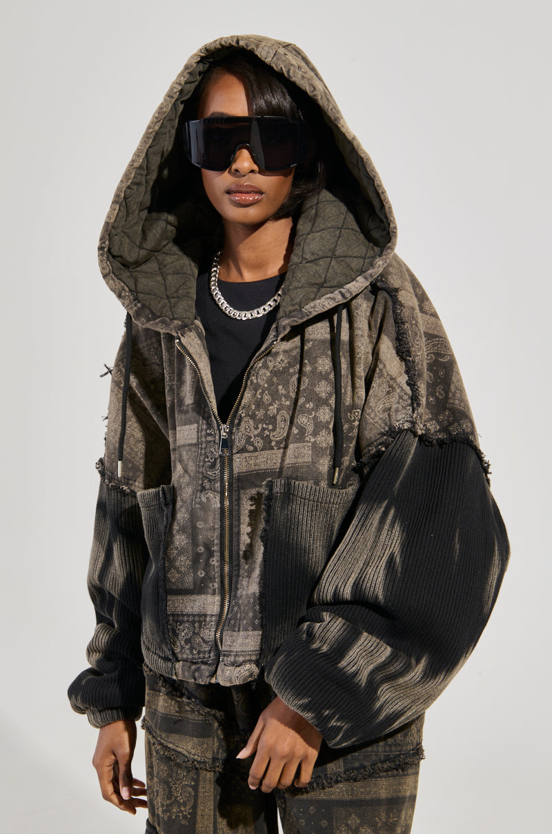 A person confidently stands in a large, patterned hoodie and sunglasses. The PHOENIX HOODED BOMBER features a paisley print and various patches, enhanced by its cotton knit fabrication. A chain necklace completes the ensemble against a plain, neutral background.
