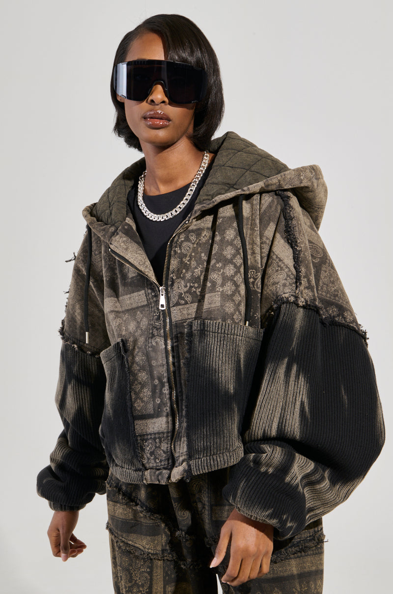 A person wearing oversized black sunglasses and a Phoenix Hooded Bomber paired with paisley print matching pants. The outfit showcases cotton knit fabrication for a blend of textures and colors. They also accessorize with a silver chain necklace, set against a plain, light-colored background.