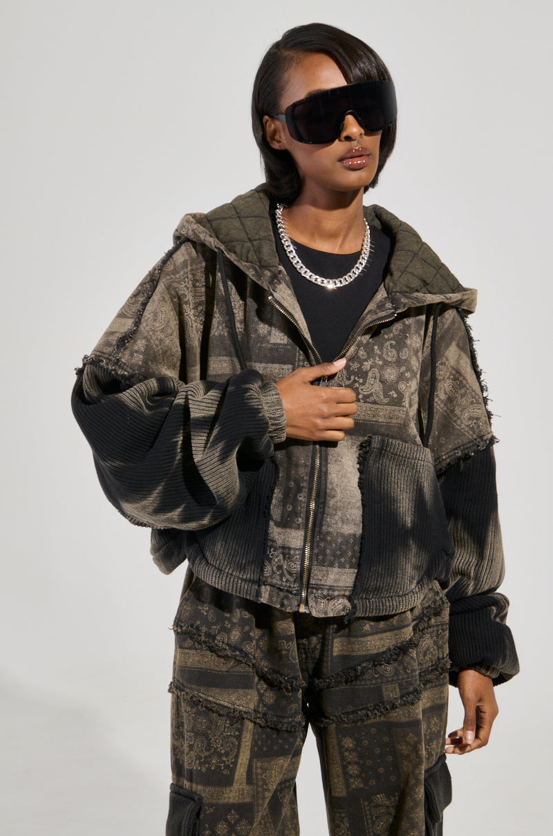 A person wears oversized sunglasses and a dark patterned Phoenix Hooded Bomber with matching pants. They have short hair and a layered necklace. The clothing features various textures, including cotton knit fabrication, creating a stylish, contemporary look with subtle paisley print accents.