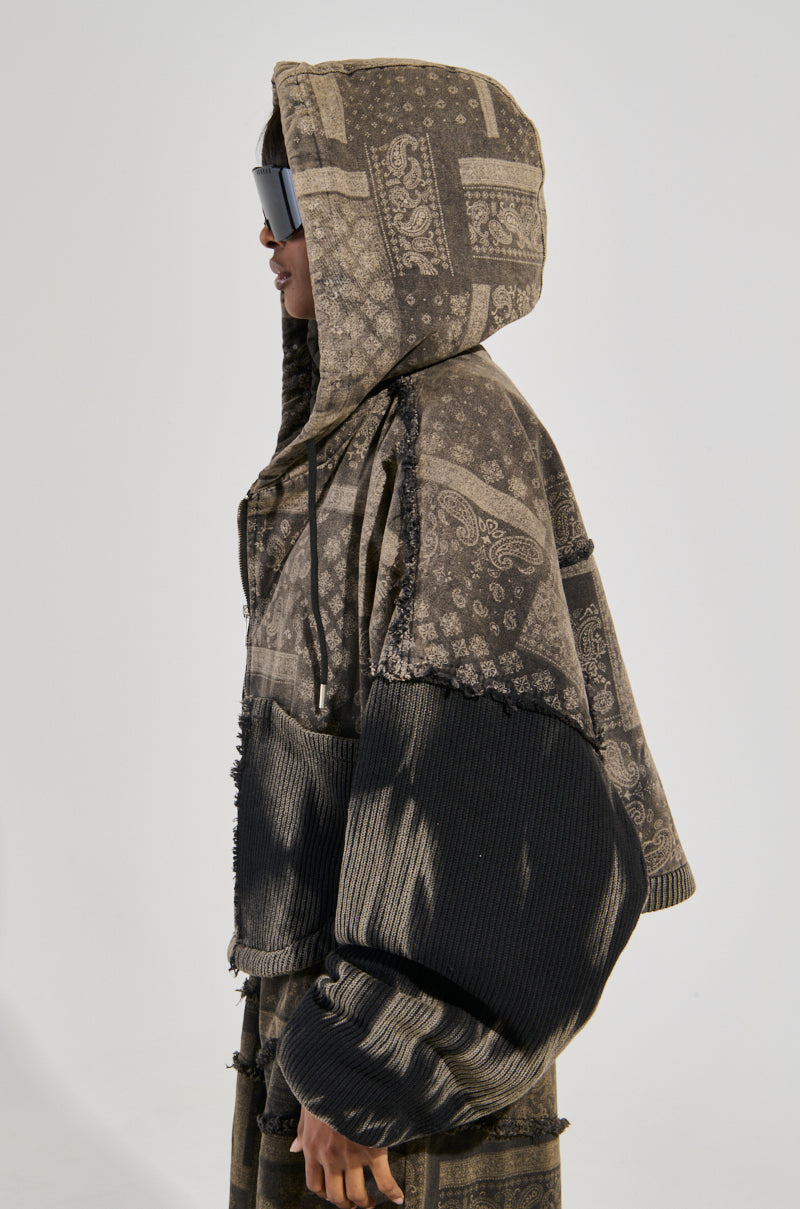 A person wearing the PHOENIX HOODED BOMBER, featuring a patchwork design in earthy tones, stands sideways. The cotton knit fabrication provides comfort, while ribbed, oversized sleeves and sunglasses complete the look. The paisley print subtly enhances the jacket's unique style.