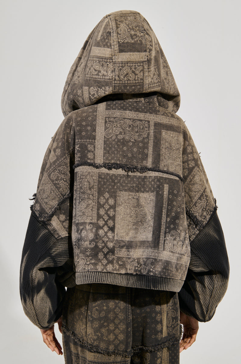 A person wearing a "Phoenix Hooded Bomber" with patchwork-style paisley print in muted tones of gray and beige stands facing away. The cozy cotton knit fabrication pairs perfectly with matching pants, creating a cohesive outfit against a plain light background.