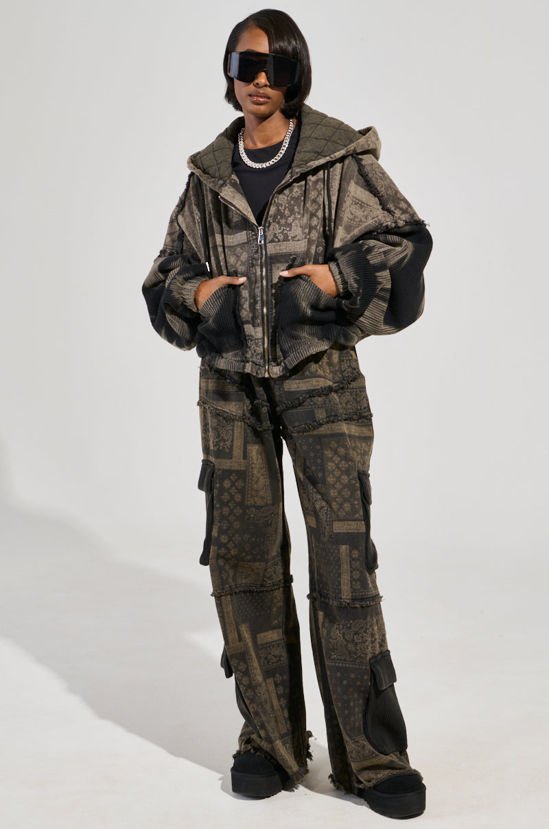 A person in a stylish, oversized gray and black jacket with a hood and matching paisley print pants exudes confidence. The PHOENIX HOODED BOMBER features cotton knit fabrication, while their short dark hair, sunglasses, and chain necklace complete the look against a neutral background.