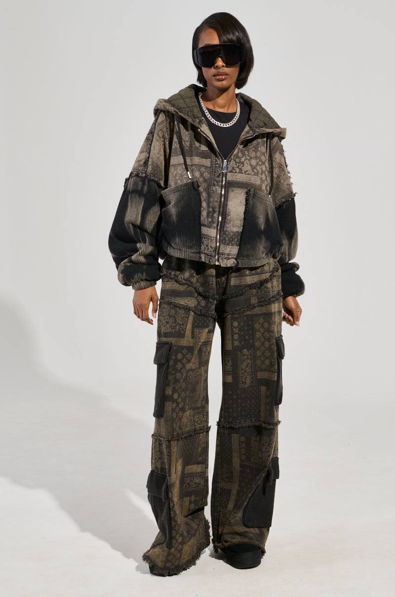 A person confidently stands against a plain background, wearing oversized sunglasses paired with the PHOENIX HOODED BOMBER, a distressed cotton knit jacket. They complete their look with matching wide-leg pants featuring patchwork patterns. The dark-toned outfit is accented by a hint of paisley print, bringing an eclectic touch to the mix of textures.